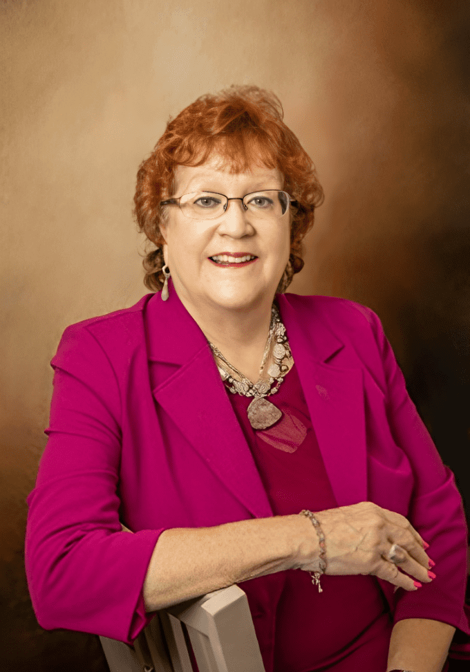 A woman with red hair and glasses wearing a pink jacket.